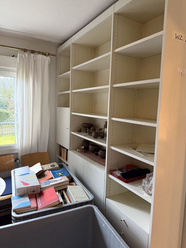 Bücherregal in Bergisch Gladbach fast ausgeräumt Ein weißes Regal, das bis auf ein paar Gegenstände leer ist, davor ein grauer Rollbehälter voller Bücher