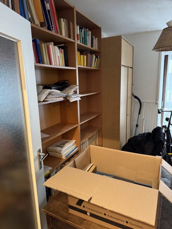 Ein volles Bücherregal mit Papieren, davor steht ein offener Umzugskarton bereit