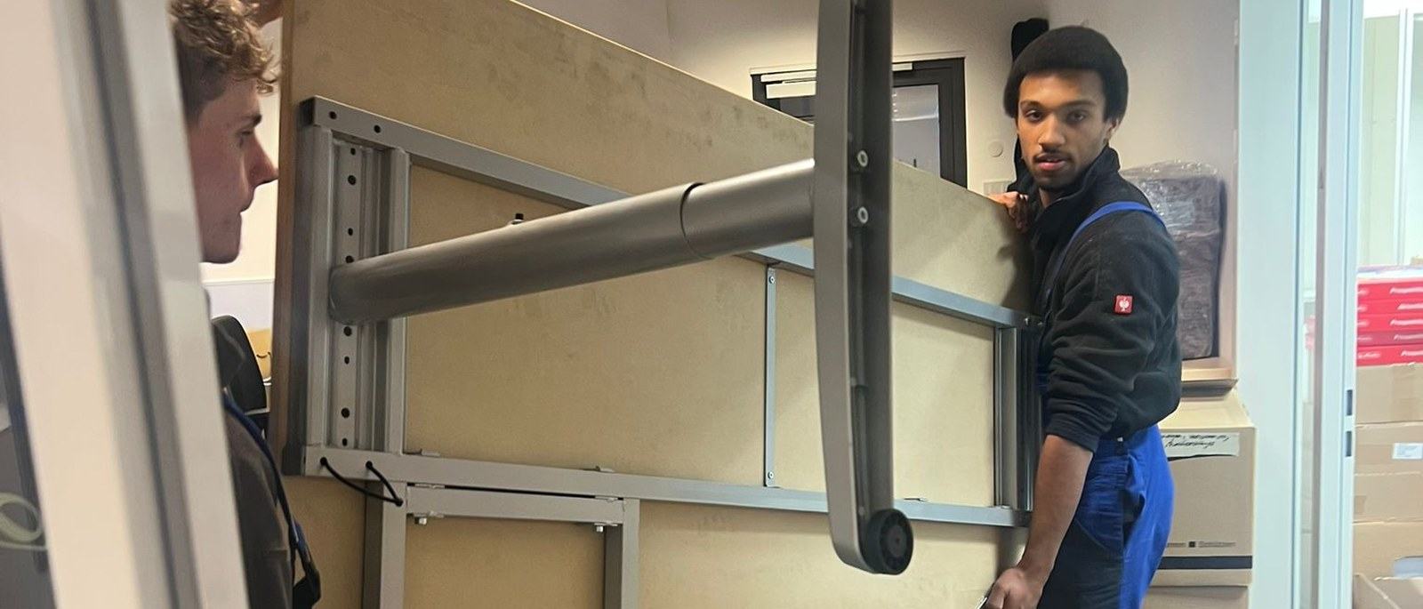 Büroumzug in Bergisch Gladbach: Zwei Möbelpacker bringen einen aufrecht gekippten Schreibtisch durch die Bürotür, hinter ihnen stapeln sich gepackte Umzugskartons