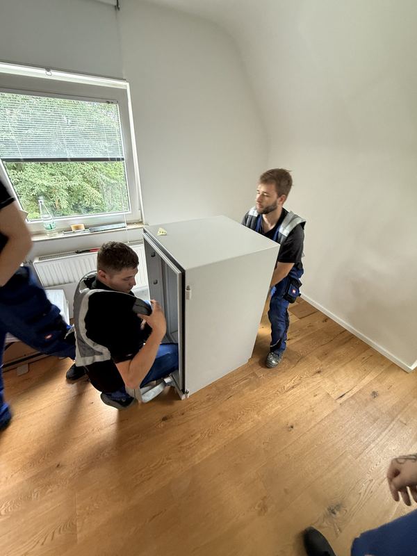 Zwei Kollegen tragen ein weißes Einbaugerät durch einen leeren Raum