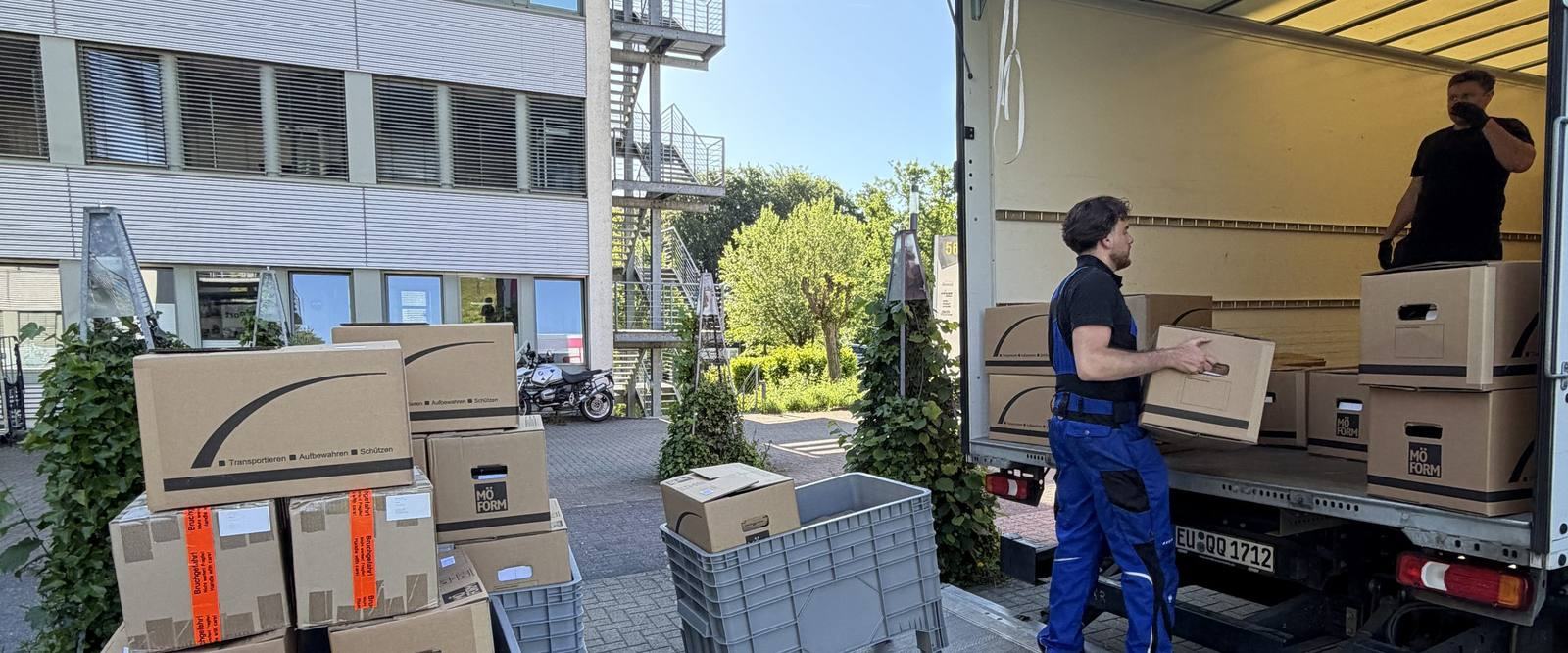 Unser Umzugsalltag in Bergisch Gladbach in Bildern: Vor einem Bürogebäude mit Außentreppe steht ein Umzugswagen mit offenem Laderaum, ein Mitarbeiter trägt einen Karton zur Ladebordwand, ein zweiter steht im Laderaum, im Vordergrund gestapelte Umzugskartons und graue Kunststoffboxen.