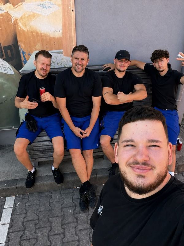 Fünf Kollegen in Arbeitskleidung sitzen nebeneinander auf einer Bank in der Sonne