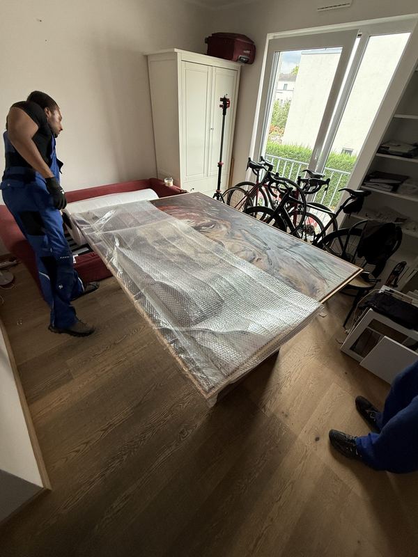 Bild mit Rahmen in Luftpolsterfolie Auf einem Tisch liegt ein großes gerahmtes Bild, eingeschlagen in Luftpolsterfolie, dahinter ein Mitarbeiter in einem Raum mit Fahrrädern