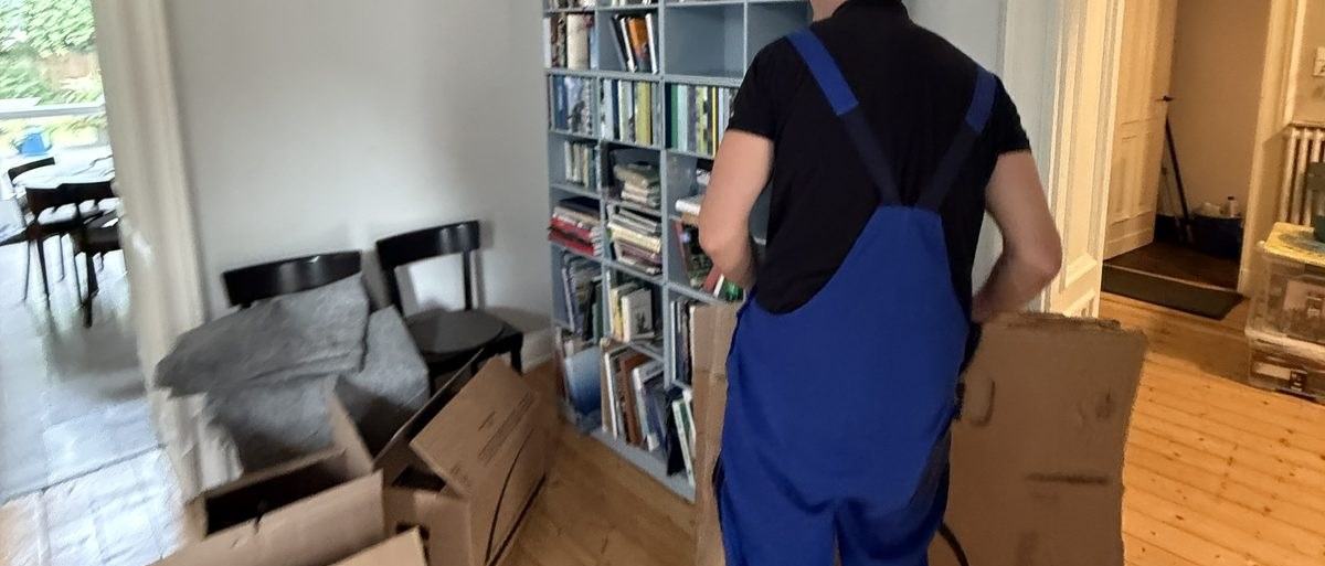 Haushaltsauflösung in Bergisch Gladbach: Ein Mitarbeiter in Latzhose geht mit einem gefalteten Karton an einem gut gefüllten Bücherregal vorbei, vor ihm Stühle und gestapelte Kartons