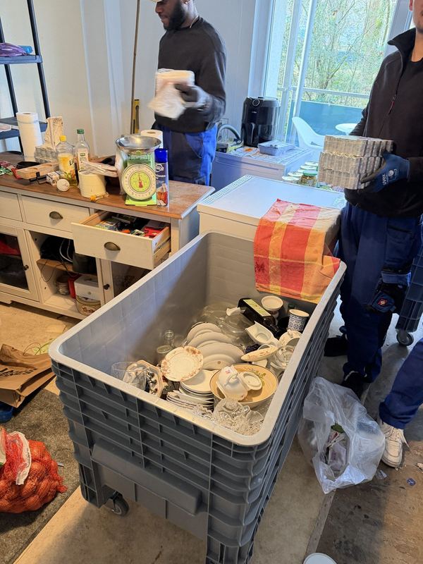Geschirr im Rollbehälter In einer Küche steht ein grauer Rollbehälter, gefüllt mit Tellern und Geschirr, im Hintergrund sortieren zwei Mitarbeiter den restlichen Hausrat
