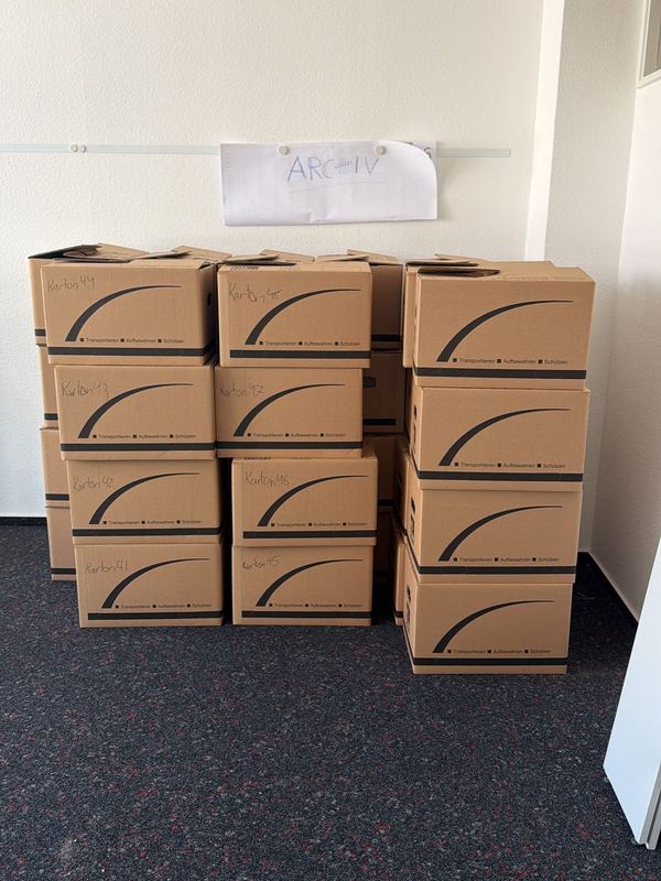 Kartons nach Zielraum sortiert Gestapelte Umzugskartons mit handschriftlicher Beschriftung in einem ausgeräumten Zimmer mit dunklem Teppich, an der Wand klebt ein Zettel, auf dem Archiv steht