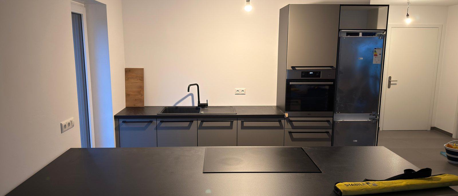 Küchenmontage in Bergisch Gladbach: Eine fertig montierte moderne Küche mit dunklen Fronten, Kochinsel mit Induktionsfeld, Spüle, Backofen und Kühlschrank in einem hellen leeren Raum, auf dem Boden liegt noch eine Werkzeugtasche.