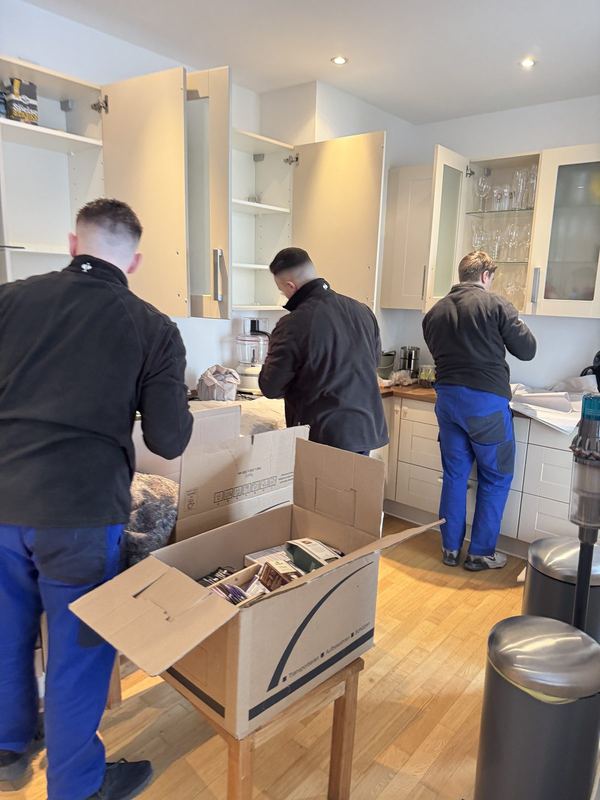 Küchenschränke leeren und verpacken In einer weißen Küche leeren drei Mitarbeiter Ober- und Unterschränke und packen den Inhalt auf einem Tisch in Kartons