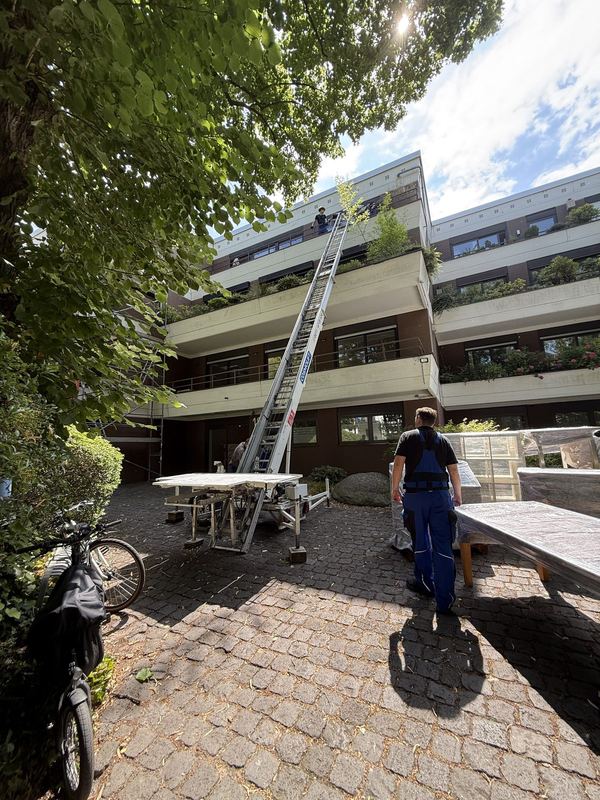 Lift bis zum Balkon Am Balkon eines mehrstöckigen Wohnhauses lehnt ein ausgefahrener Möbellift, vorne auf einem gepflasterten Weg stehen ein Mitarbeiter und Tischplatten