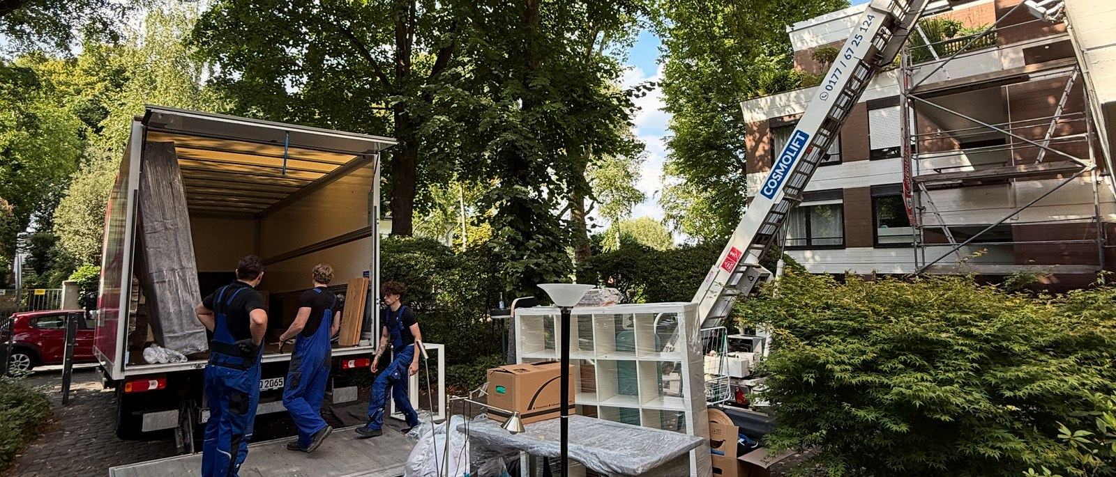 Möbellift für Umzüge in Bergisch Gladbach: Ein Möbellift ist an einem eingerüsteten Wohnhaus bis zum obersten Geschoss ausgefahren, drei Mitarbeiter stehen vor dem geöffneten Laderaum eines Lkw neben einem Regal und Kartons