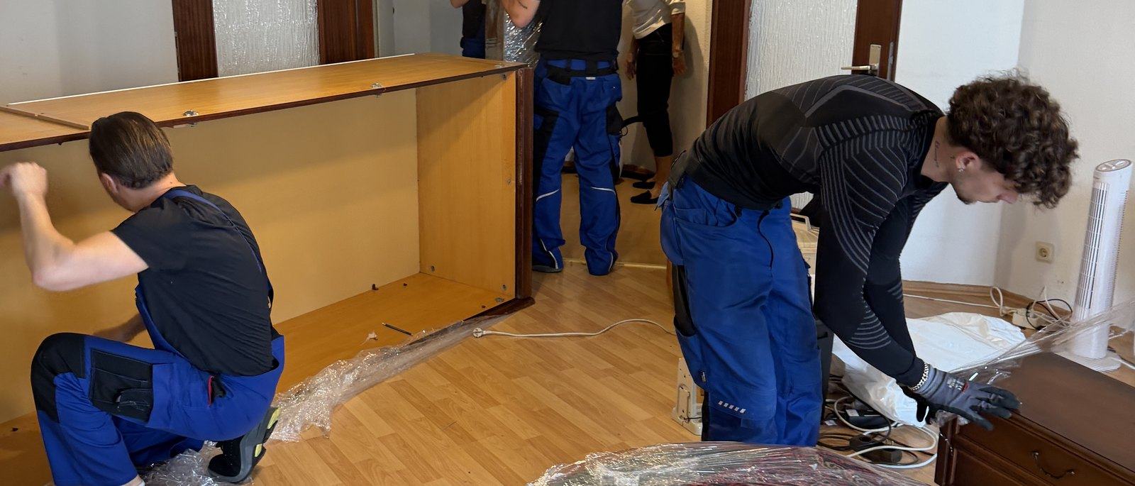 Möbelmontage in Bergisch Gladbach: Drei Mitarbeiter zerlegen in einem Wohnzimmer mit Parkett eine große Schrankwand aus Holz, die Seitenteile liegen bereits am Boden, daneben Folie und Werkzeug.