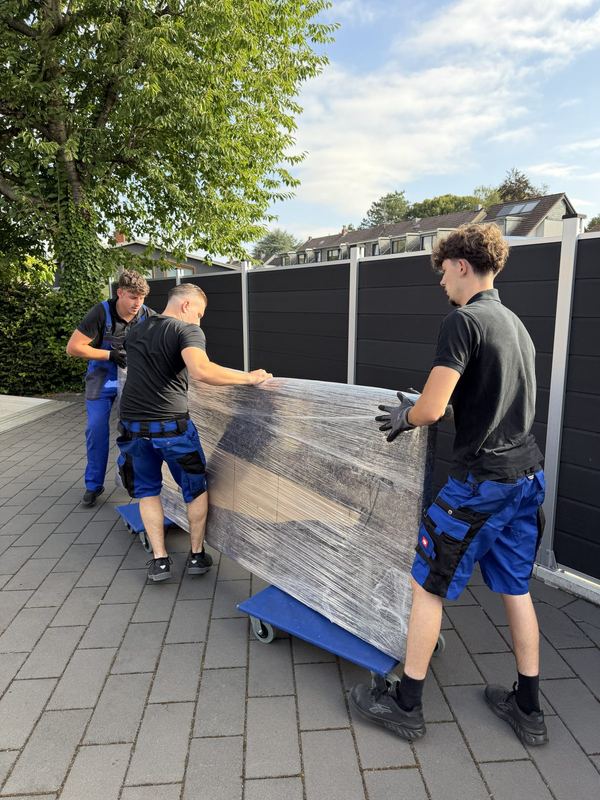 Drei Kollegen rollen ein foliertes Möbelstück auf Rollbrettern über eine Einfahrt