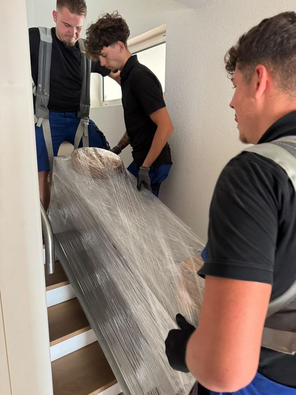 Drei Kollegen tragen zusammen ein in Folie gewickeltes Möbelstück die Treppe hinab