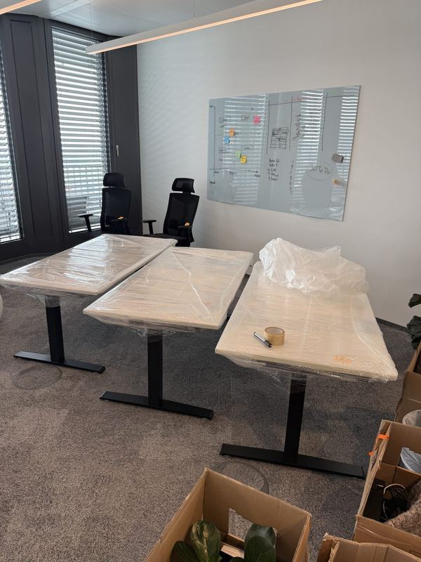 Folierte Schreibtischplatten Drei höhenverstellbare Schreibtische, deren Platten in Stretchfolie gewickelt sind, stehen in einem Besprechungsraum vor Whiteboard und Bürostühlen