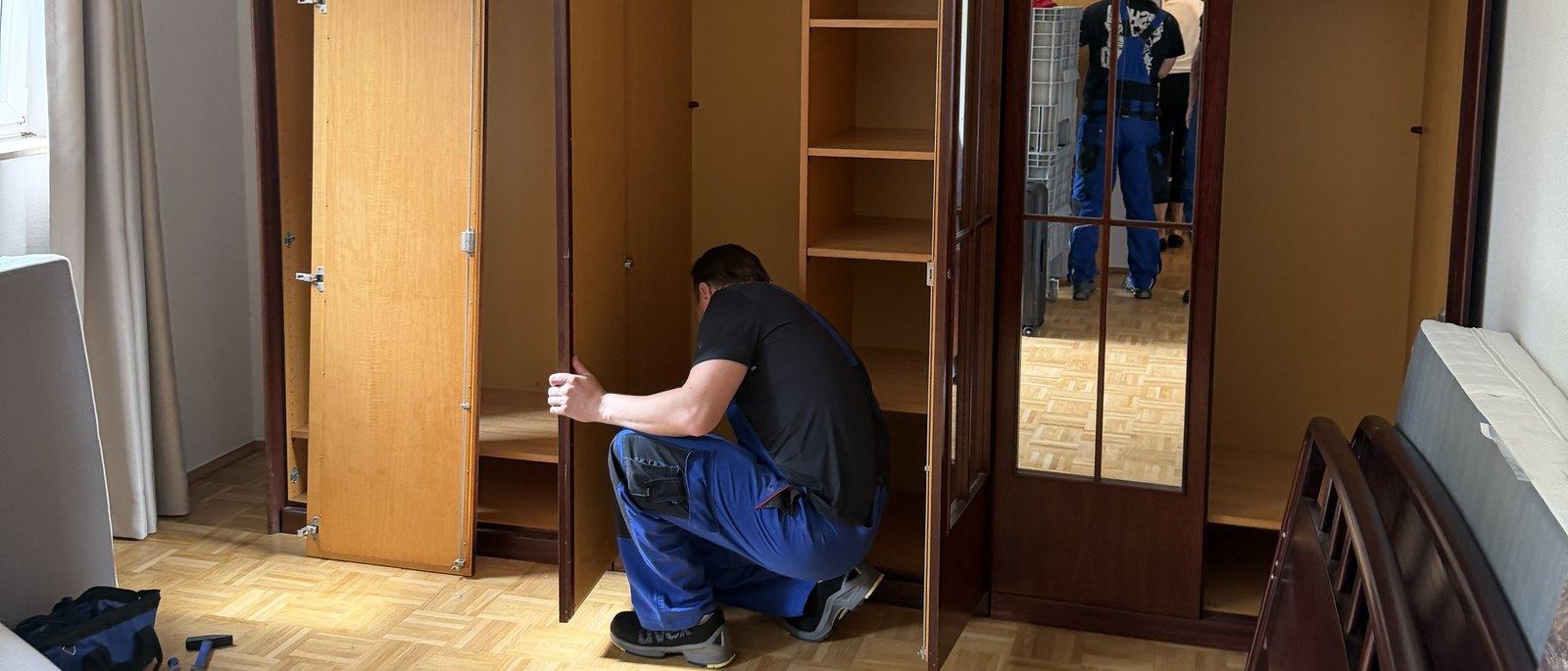 Seniorenumzug in Bergisch Gladbach: In einem Schlafzimmer mit Parkett kniet ein Mitarbeiter vor einem leer geräumten Kleiderschrank aus dunklem Holz