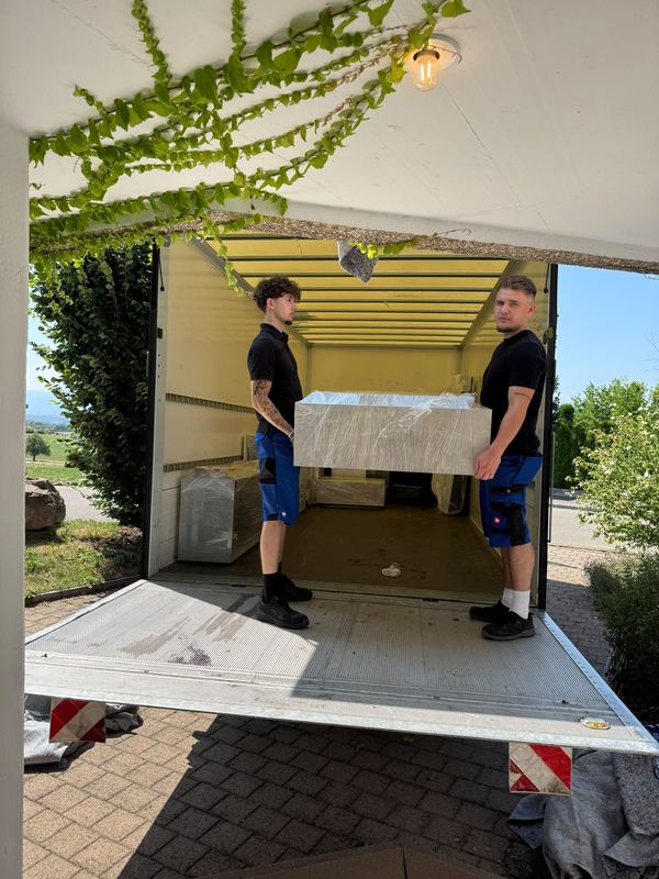 Verpacktes Möbelstück über die Ladebordwand in den Wagen Zwei Kollegen heben ein foliertes Möbelstück über die Ladebordwand hinein in den Laderaum des Umzugswagens