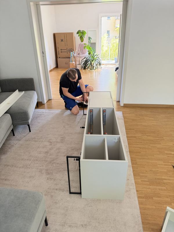 Aufbau des Sideboards in der neuen Wohnung Ein Kollege kniet vor einem weißen Sideboard in einem lichtdurchfluteten Wohnzimmer und baut es zusammen, dahinter stapeln sich Umzugskartons
