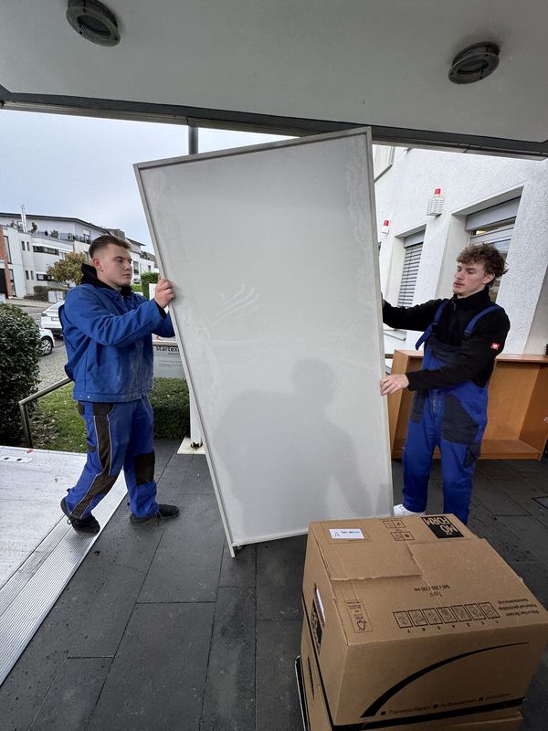 Zwei Kollegen tragen eine große weiße Platte hochkant zum Wagen