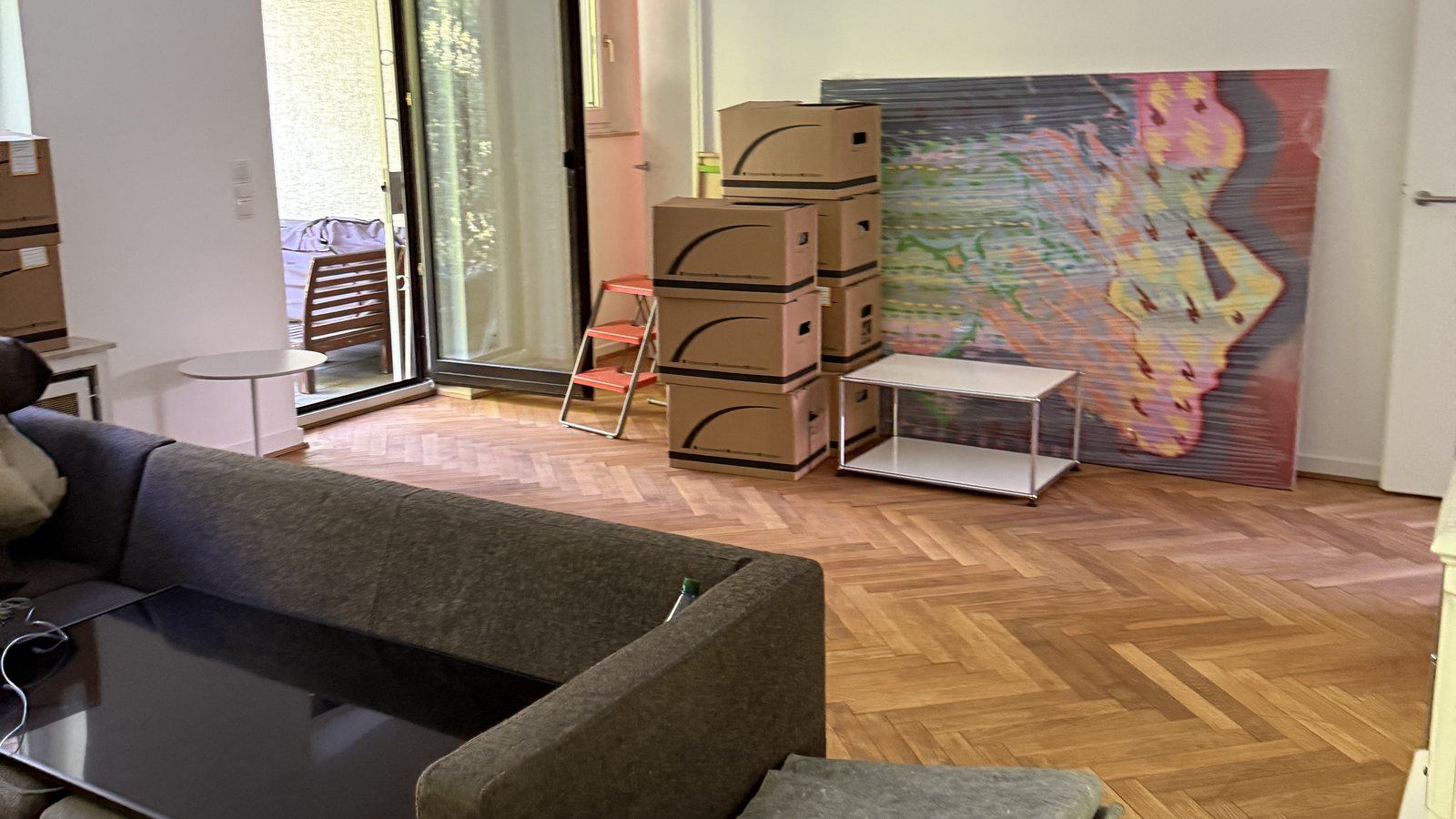 Umzugscheckliste für Bergisch Gladbach: In Etappen ins neue Zuhause: In einem Wohnzimmer mit Fischgrätparkett stehen gestapelte Umzugskartons neben einem Sofa und einem Beistelltisch, an der Wand lehnt ein großes buntes Bild, dahinter eine Terrassentür.