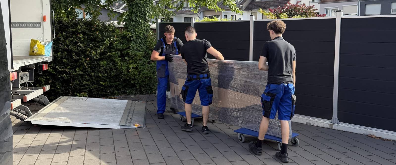 Umzugsunternehmen Bergisch Gladbach: Drei Kollegen schieben ein folienverpacktes Möbelstück auf Rollbrettern zum Umzugswagen, dessen Ladebordwand abgesenkt ist
