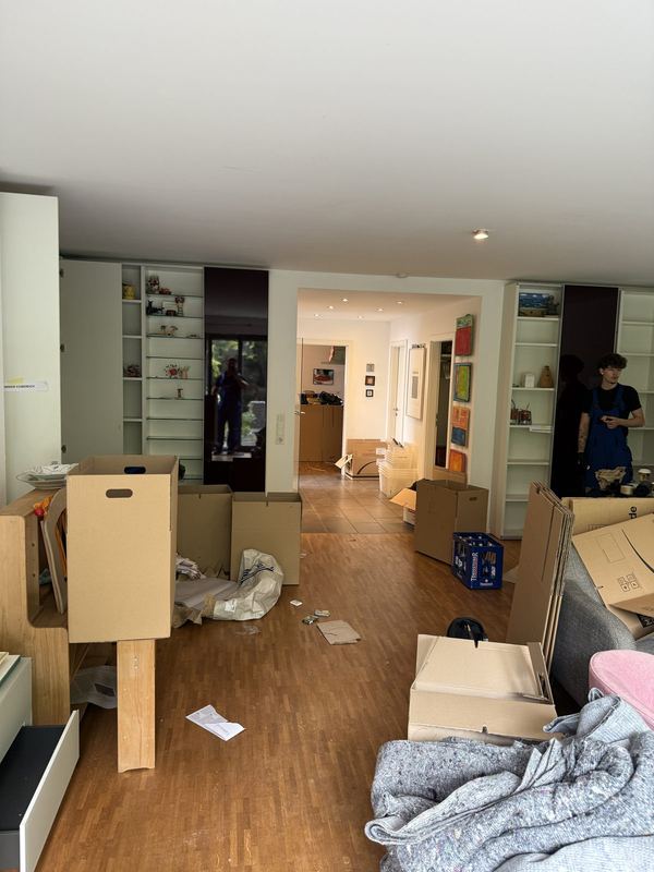 Ein Wohnzimmer mit schon leeren Einbauregalen und aufgestellten Kartons, dahinter zwei Kollegen