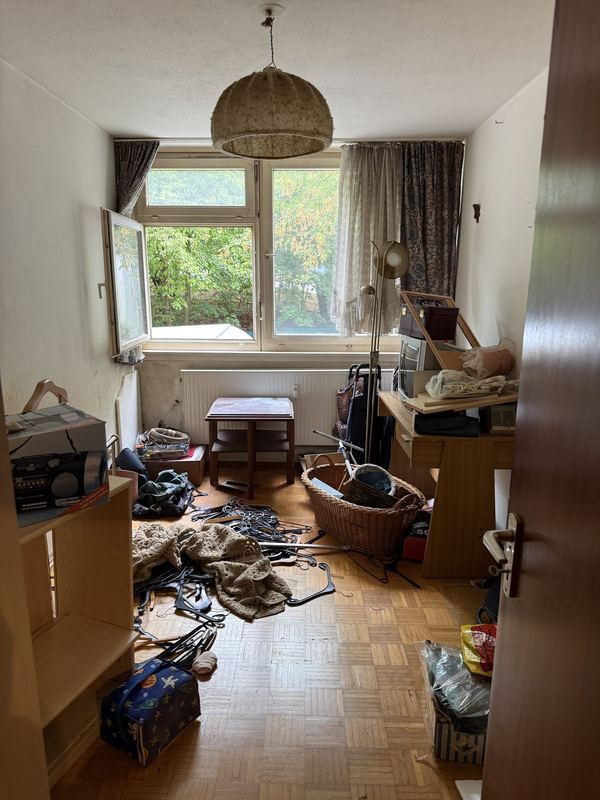 Raum vor dem Ausräumen Ein zugestelltes Zimmer mit Parkettboden, überall liegen Körbe, Kabel, Taschen und weiterer Hausrat, im Hintergrund öffnet ein Fenster den Blick ins Grüne
