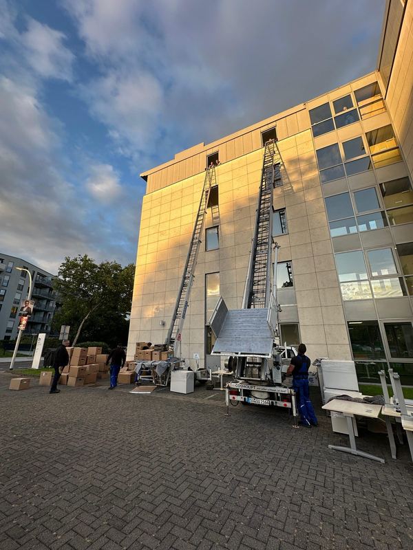 Zwei Lifte am Bürohaus An der verglasten Fassade eines Bürogebäudes stehen zwei ausgefahrene Möbellifte nebeneinander, davor ein Anhänger, Kartons und zwei Mitarbeiter