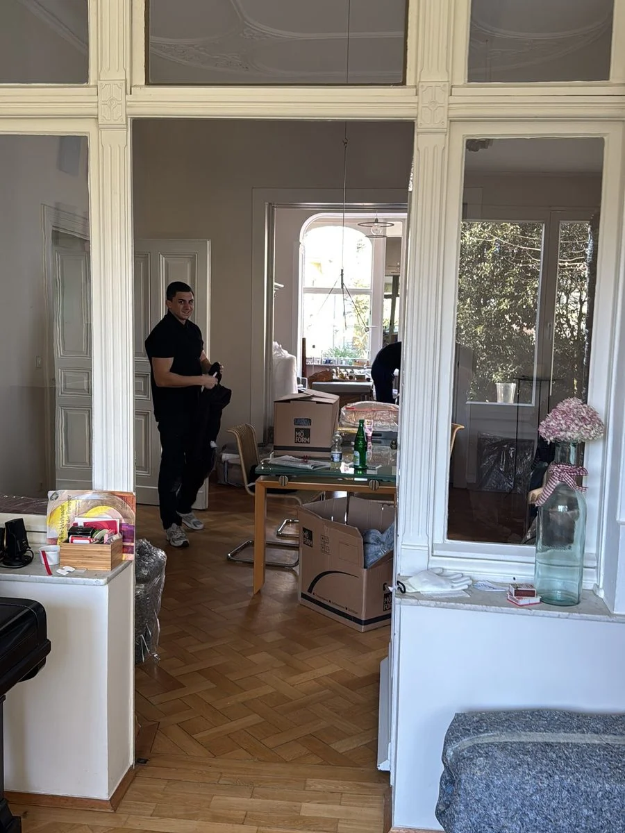 Ein Altbauzimmer mit hohen Fenstern, im Hintergrund ist klein ein Umzugshelfer bei der Arbeit zu sehen.