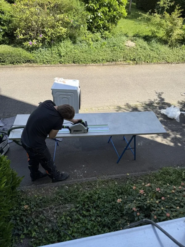 Ein Umzugshelfer schneidet mit einer Tauchsäge auf einer Führungsschiene eine Arbeitsplatte zu, die auf Böcken am Straßenrand liegt, daneben steht eine Waschmaschine auf dem Gehweg
