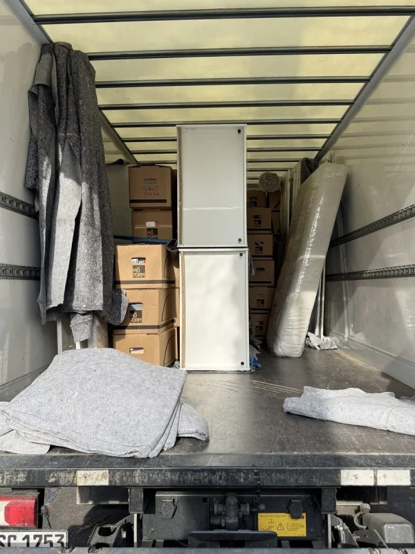 Blick von der Ladebordwand in den Laderaum eines Umzugswagens mit Kartons, einem weißen Schrank, einer gerollten Matratze und Umzugsdecken an der Wand
