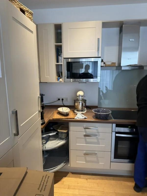 Eine helle Landhausküche mit Kassettenfronten, der Eckschrank mit Drehrondell ist geöffnet, auf der Arbeitsplatte stehen eine Küchenmaschine und Schüsseln, rechts ein Umzugshelfer
