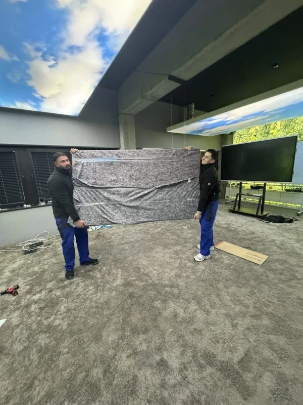 In einem Raum mit Himmelsmotiv an der Decke und einem großen Bildschirm halten zwei Umzugshelfer ein großes flaches Teil, das in eine Umzugsdecke gehüllt ist