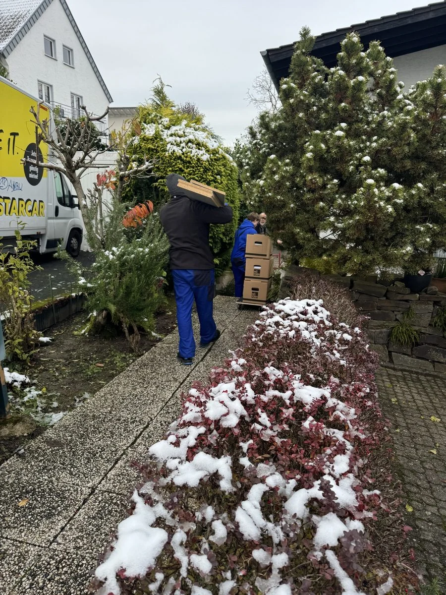 Ein Umzugshelfer trägt einen Karton durch einen verschneiten Garten in Richtung des Hauses.