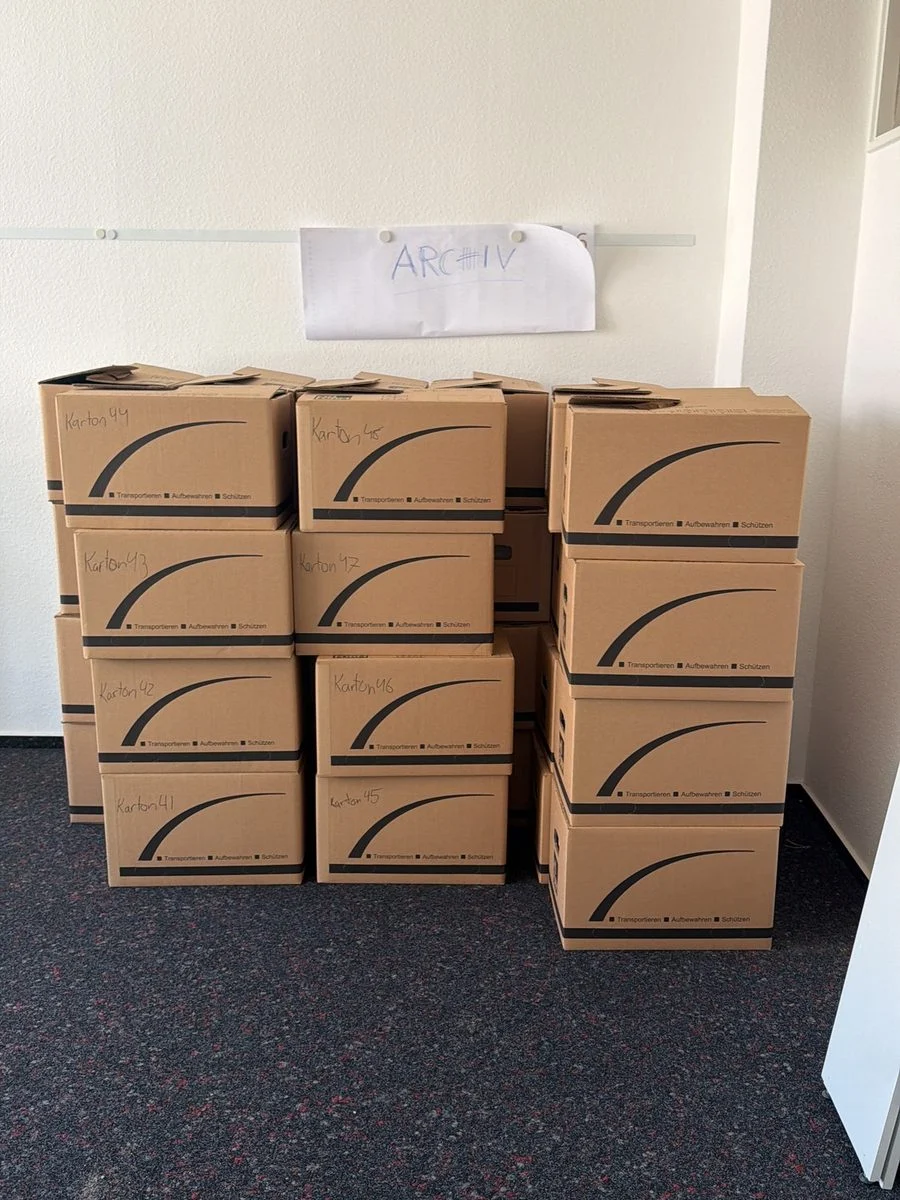 Gestapelte Umzugskartons stehen in einem Altbauzimmer, dessen Decke mit Stuck verziert ist.