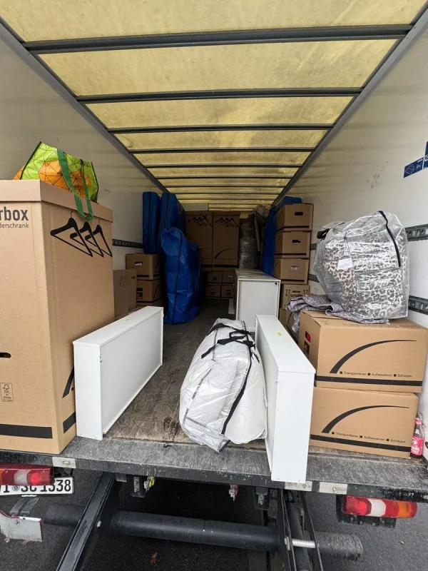 Der Laderaum eines Umzugswagens ist mit Umzugskartons, einer Kleiderbox mit Bügeln, blauen Säcken, weißen Regalböden und Taschen gefüllt