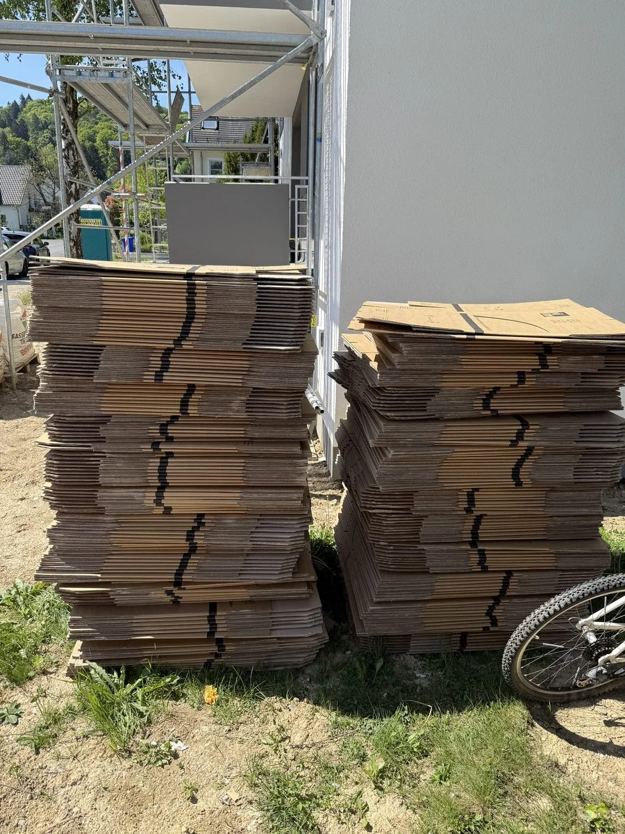 Gestapelte Umzugskartons stehen im Außenbereich vor einem Baugerüst, das an der Hausfassade aufgebaut ist.