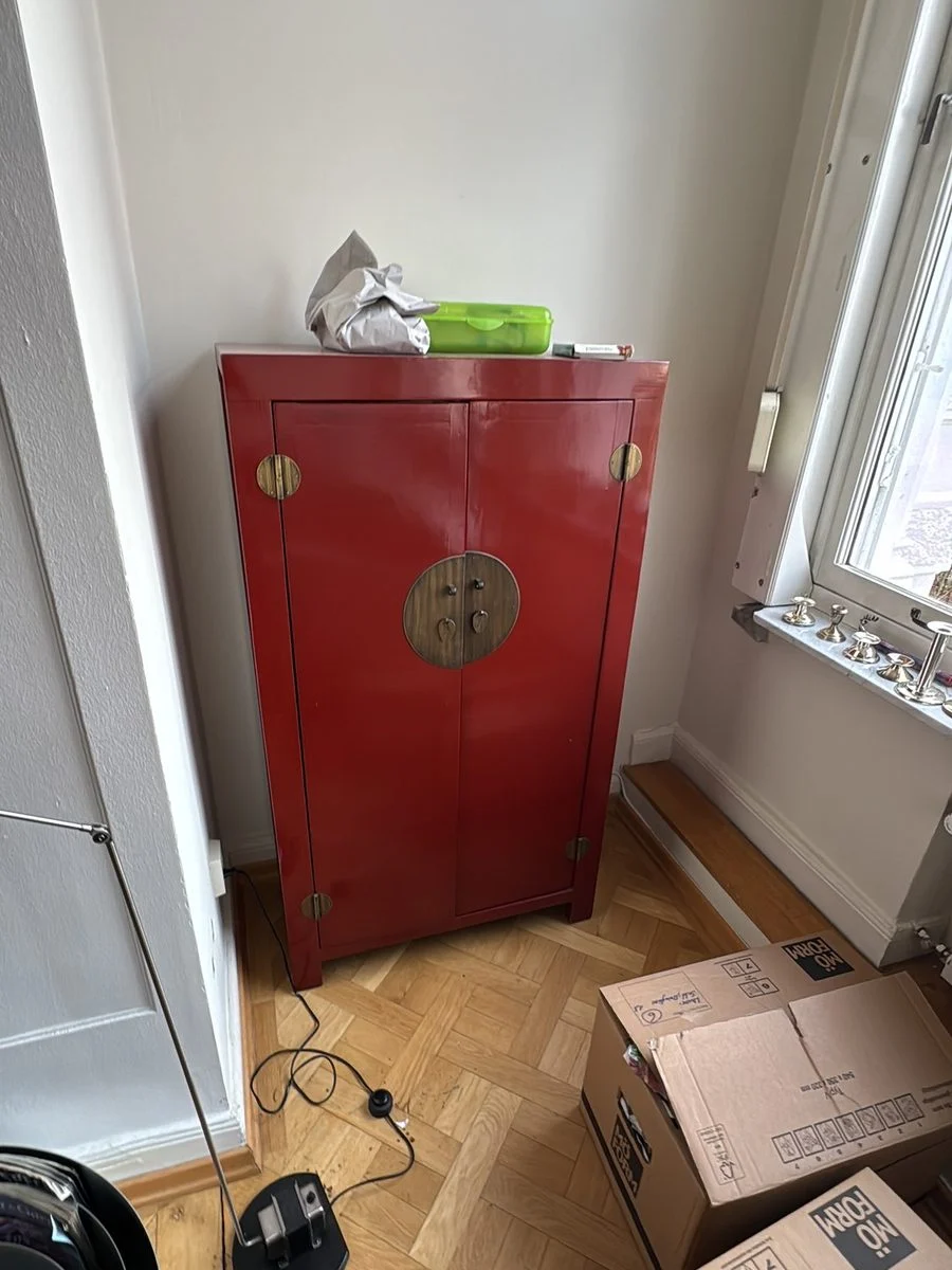 Ein roter antiker Lackschrank steht in einem Altbauzimmer, davor sind gepackte Kartons abgestellt.