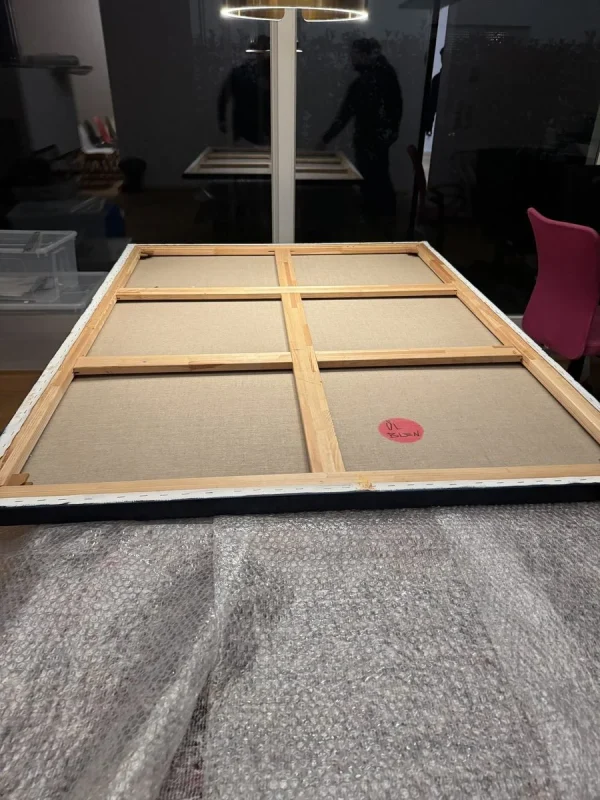 Ein großes Leinwandbild auf Keilrahmen liegt mit der Rückseite nach oben auf einer grauen Umzugsdecke, hinten spiegelt sich der Raum in einer dunklen Fensterfront