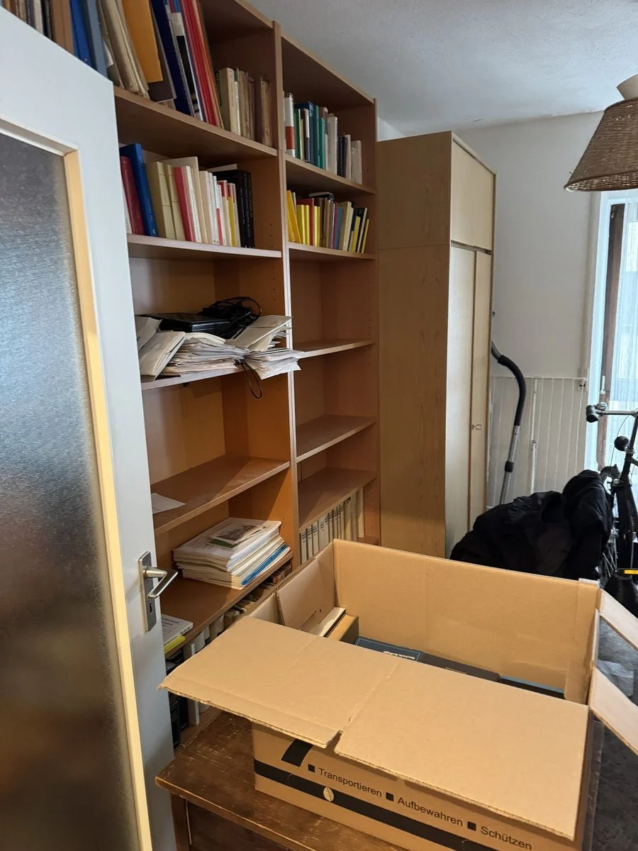 Vor einer Regalwand steht ein geöffneter, leicht unaufgeräumter Umzugskarton auf dem Boden.