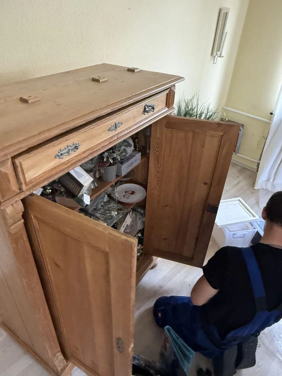 Ein Umzugshelfer kniet vor einem geöffneten antiken Schrank und ist mit der Demontage beschäftigt.