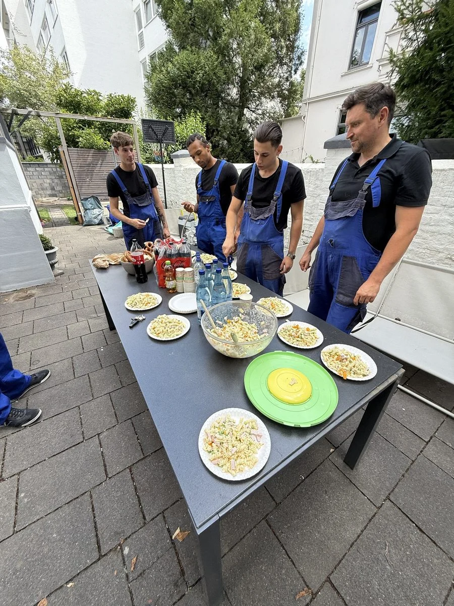 Das Team in Arbeitskleidung sitzt an einem Tisch im Freien und nimmt gemeinsam eine Mahlzeit ein.