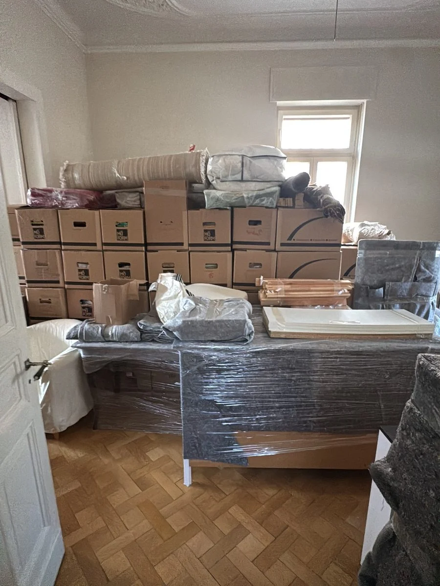 In einem Zimmer stehen gestapelte Umzugskartons und in Folie verpackte Möbel auf dem Parkettboden.