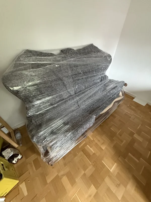 In einer Zimmerecke auf Parkett steht ein Sofa, das in eine graue Umzugsdecke und Stretchfolie eingepackt ist