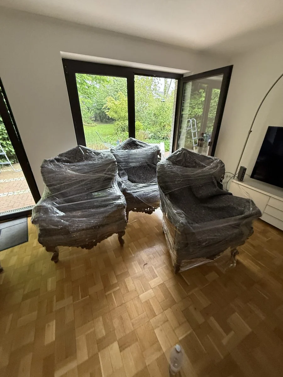 In einem Wohnzimmer stehen vier verpackte Sessel im Kreis vor großen Fenstern.