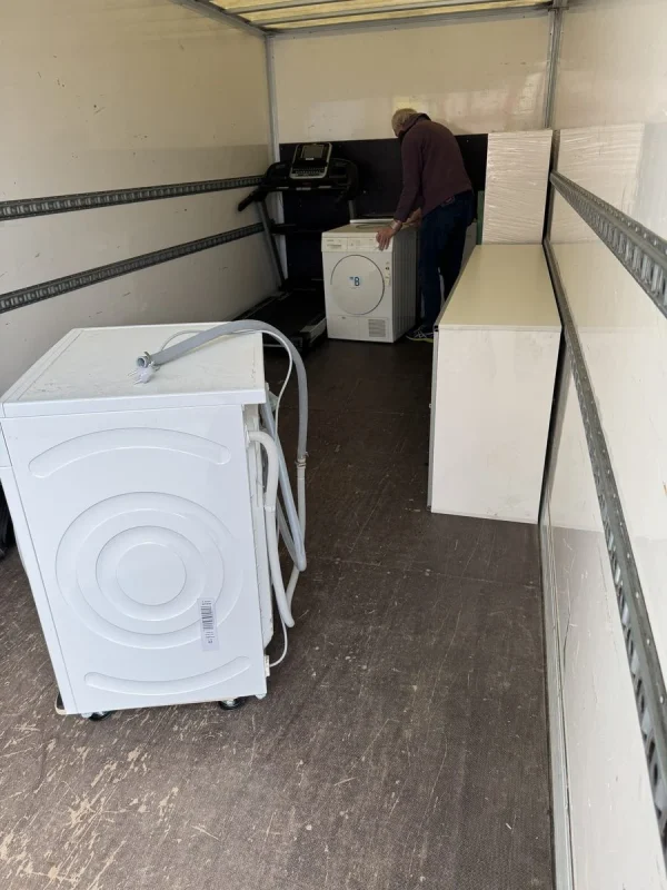 Eine Waschmaschine steht auf einem Rollbrett im Laderaum eines Umzugswagens neben weißen Möbeln, im Hintergrund sichert ein Umzugshelfer ein weiteres Gerät