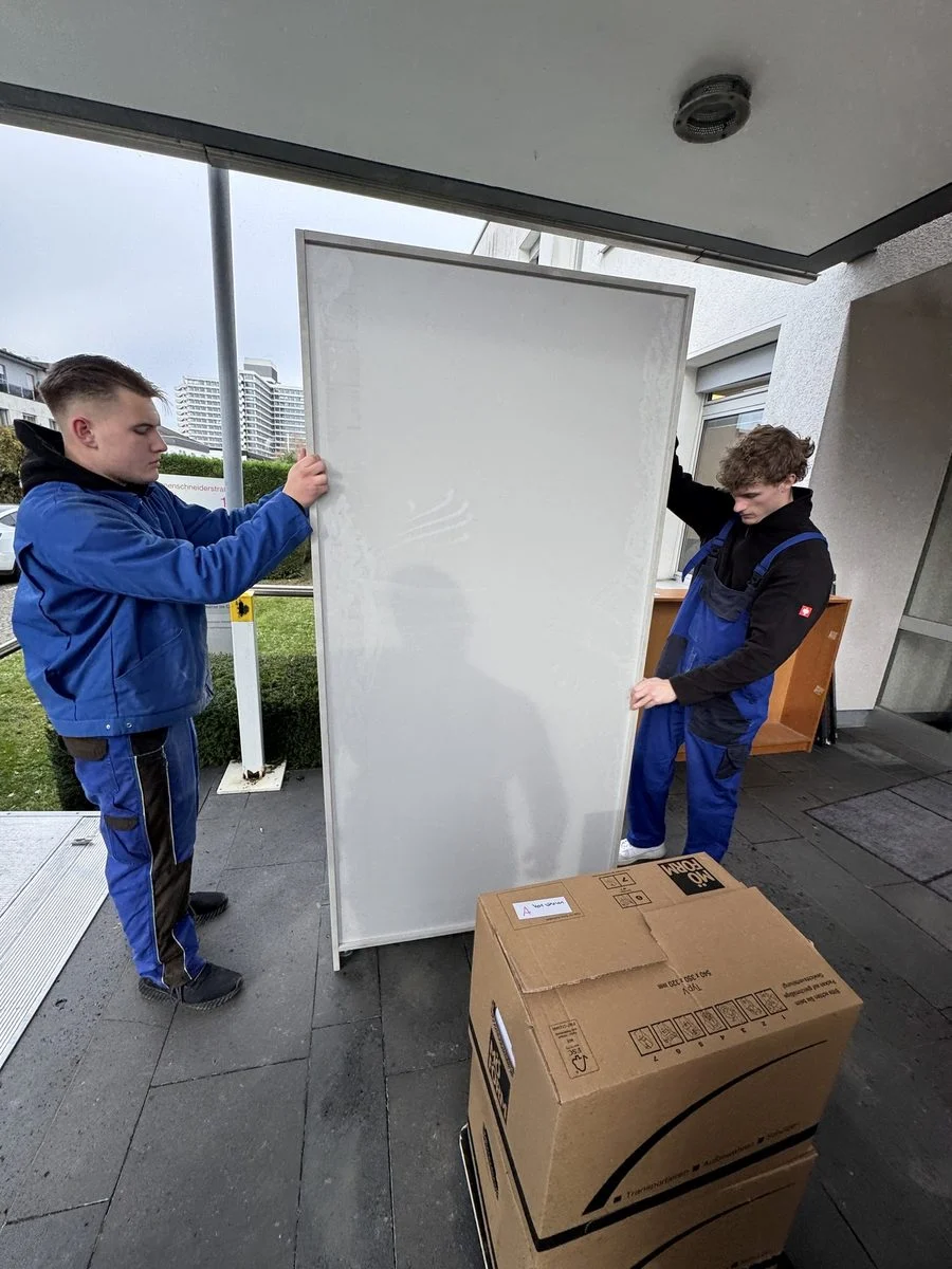 Zwei Kollegen tragen gemeinsam eine große weiße Tür hinaus auf einen Balkon.