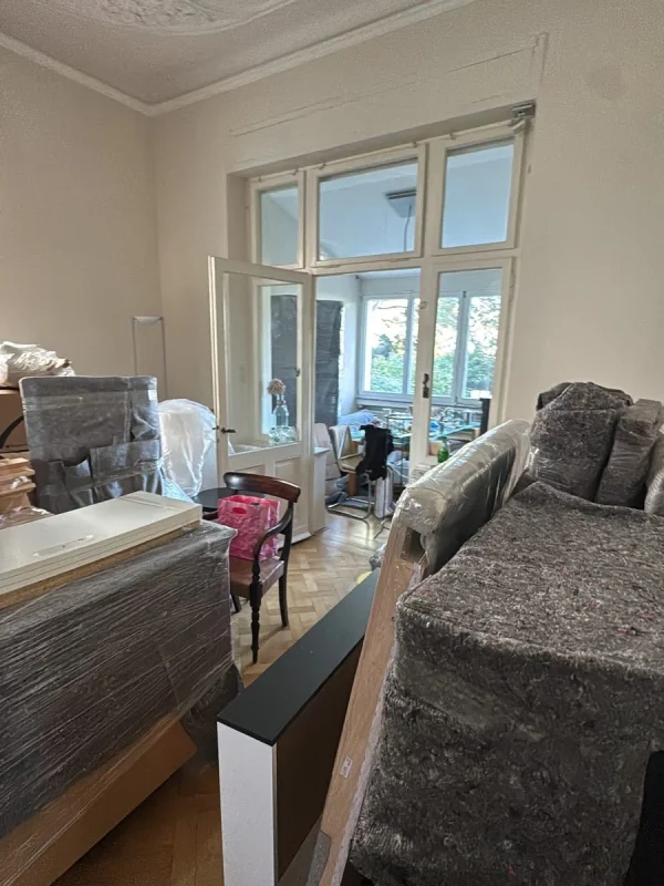 In einem Altbauwohnzimmer mit Stuck und Flügeltür stehen Möbel in Umzugsdecken und Folie eingepackt, dazwischen mehrere Stühle
