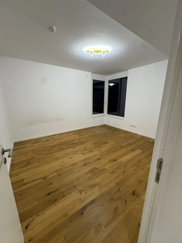 Ein leeres Zimmer mit Holzdielen und weißen Wänden, die Deckenlampe brennt, hinter dem Fenster ist es bereits dunkel